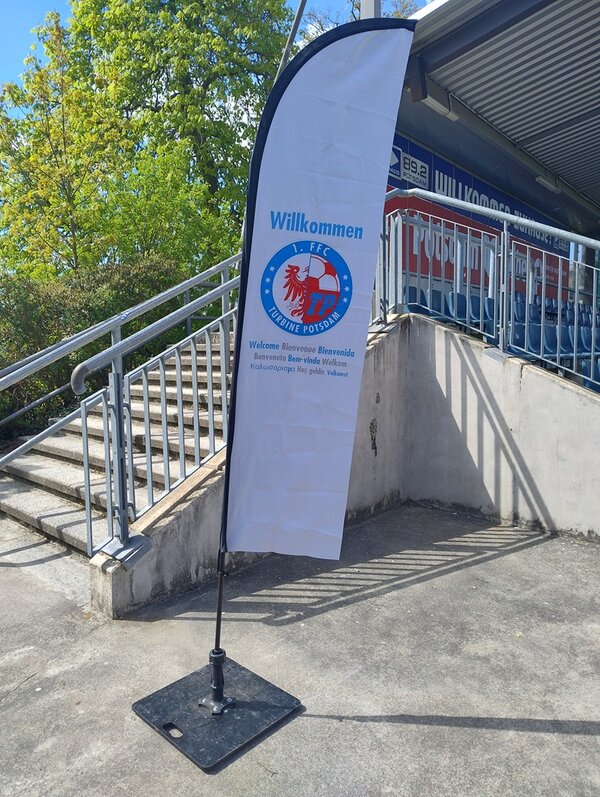 Ein hochformatiges, weißes Banner mit dem Logo und Schriftzug eines Fußballvereins steht auf einem schwarzen Standfuß vor einer Metalltreppe im Außenbereich eines Stadions. Das Banner zeigt oben das Wort 'Willkommen', darunter das runde Vereinslogo mit einem roten Greif und blauen Elementen sowie weiteren Text. Im Hintergrund sind Tribünen mit blauen Sitzen und ein Teil eines Stadiondachs zu sehen. Rechts wachsen grüne Bäume, die im Sonnenlicht leuchten.