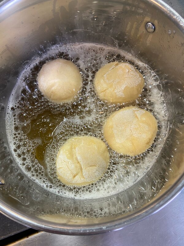 Mehrere ungebackene Krapfen-Teiglinge werden in einem heißen Fettbad in einem Edelstahltopf frittiert. Blasen steigen an die Oberfläche, während der Teig langsam goldbraun wird.