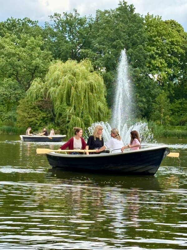 Zwei Gruppen von Menschen sitzen in Ruderbooten auf einem See und genießen die Zeit auf dem Wasser. Im Hintergrund ist ein großer Springbrunnen zu sehen, der Wasser in die Luft spritzt. Um den See herum sind Bäume und grüne Landschaft zu sehen.