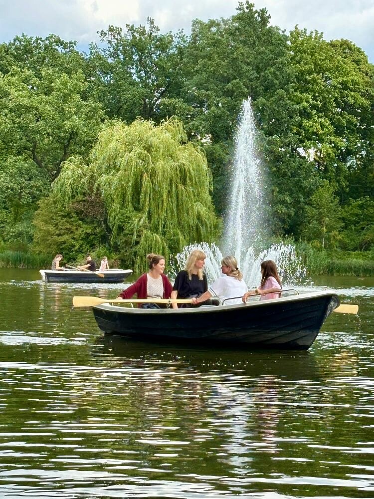 Zwei Gruppen von Menschen sitzen in Ruderbooten auf einem See und genießen die Zeit auf dem Wasser. Im Hintergrund ist ein großer Springbrunnen zu sehen, der Wasser in die Luft spritzt. Um den See herum sind Bäume und grüne Landschaft zu sehen.