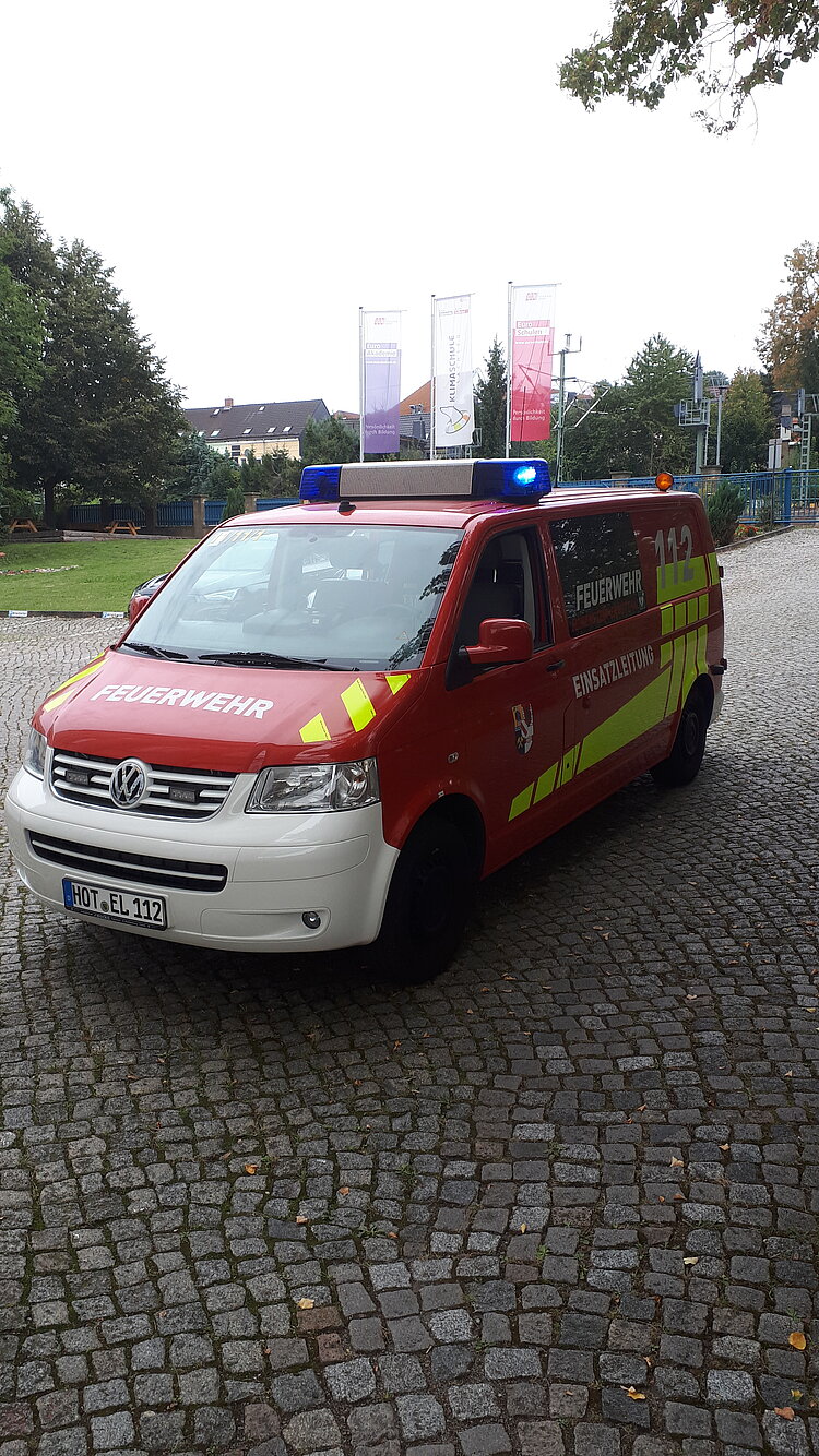 Ein rotes Einsatzfahrzeug der Feuerwehr mit gelben Streifen und der Aufschrift „Einsatzleitung“ sowie der Notrufnummer 112 steht auf einem gepflasterten Platz. Auf dem Dach sind Blaulichter eingeschaltet, im Hintergrund sind Fahnen der Euro-Schulen zu sehen.