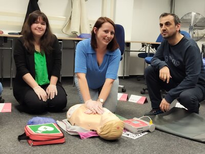 Drei Personen nehmen an einem Erste-Hilfe-Kurs teil. Eine Frau in einem blauen Pullover demonstriert die Herz-Lungen-Wiederbelebung (HLW) an einer Übungspuppe, während zwei weitere Teilnehmer – eine Frau in einer schwarzen Jacke und ein Mann in einem dunklen Pullover – daneben knien und zuschauen. Vor ihnen liegt ein Erste-Hilfe-Set mit einem Defibrillator.