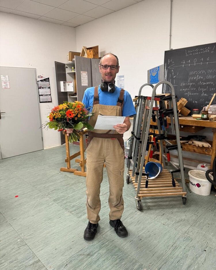 Der Mitarbeiter, der sein 20-jähriges Dienstjubiläum feiert, steht in einer Werkstatt und hält einen Blumenstrauß sowie ein Blatt Papier in den Händen. Er trägt eine beigefarbene Latzhose über einem blauen T-Shirt und hat Kopfhörer um den Hals. Im Hintergrund sind Regale mit Werkzeugen, eine Tafel mit handgeschriebenem Text und verschiedene Arbeitsmaterialien zu sehen. Der Boden ist hellgrau, die Wände weiß.
