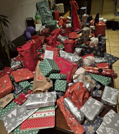 Ein großer Tisch ist vollständig mit zahlreichen Weihnachtsgeschenken bedeckt, die in verschiedenen bunten Geschenkpapieren eingewickelt sind. Die Verpackungen zeigen Muster wie Schneeflocken, Sterne, Rentiere, Tannenbäume und andere weihnachtliche Motive in Farben wie Rot, Grün, Silber, Weiß und Blau. Die Geschenke variieren in Größe und Form, einige sind rechteckig, andere zylindrisch oder unregelmäßig geformt. Im Hintergrund sind Stühle und ein Teil eines Raumes mit Fliesenboden zu sehen. Die Szene vermittelt eine festliche Atmosphäre im Rahmen der Weihnachtsfeier der Euro-Schulen Rhein-Ruhr-Wupper GmbH.
