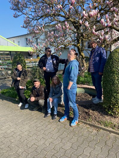 Mehrere Personen stehen und sitzen an einem sonnigen Tag unter einem blühenden Magnolienbaum auf dem Gelände eines Secondhand- oder Gartenmarkts.