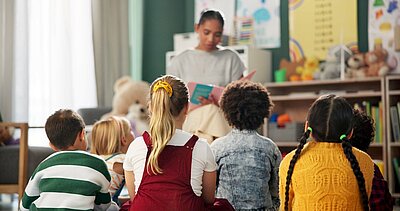 Kinder im Kindergarten sitzen nebeneinander und hören der Erzieherin zu, die aus einem Buch vorliest. 