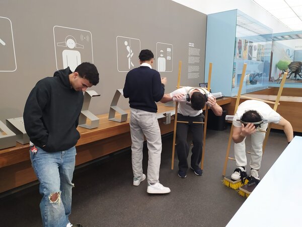 Mehrere Personen befinden sich in einem interaktiven Museum und probieren verschiedene Experimente aus. Zwei Personen haben sich in eine Vorrichtung gelehnt, die ihren Körper nach vorne kippen lässt, während eine andere Person Schuhe mit Bürsten trägt.