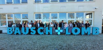 Vor einem modernen Gebäude mit großen Fenstern steht eine Gruppe von Teilnehmenden in lockerer Reihe. Im Vordergrund befindet sich ein großes, türkisfarbenes Schriftzeichen mit dem Schriftzug 'BAUSCH + LOMB'. Die Personen sind unterschiedlich gekleidet und blicken zur Kamera. Der Boden ist mit Kies bedeckt, im Hintergrund spiegeln sich Bäume in den Fenstern.