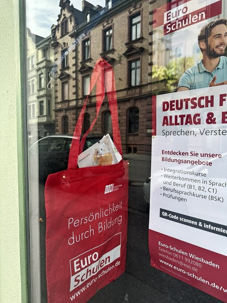 Eine rote Stofftasche mit weißem Aufdruck hängt an einem Türgriff vor einer Fensterscheibe. Auf der Tasche steht in weißer Schrift: 'Persönlichkeit durch Bildung' und darunter das Logo sowie der Name 'Euro Schulen'. Im Fenster dahinter ist ein Plakat mit weiteren Informationen zu Sprach- und Bildungsangeboten der Euro Schulen zu sehen. Die Spiegelung zeigt eine Häuserfassade mit mehreren Fenstern und Bäumen auf der gegenüberliegenden Straßenseite.