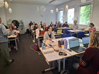 Mitarbeitende der Euro-Schulen sitzen zur Weiterbildung in einem großen Unterrichtsraum an Tischen, über ihnen hängen weiße Papiersterne, grauer Teppichboden ist ausgelegt.