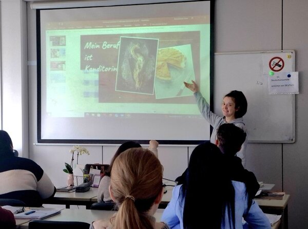 Eine weibliche Teilnehmerin steht vorn am Whiteboard und zeigt mit dem Finger auf das Whiteboard, auf dem sie ihre Präsentation vorträgt.