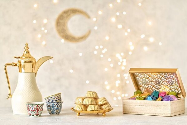 Ein Bild zum Ramadan für Muslime. Eine gold-weiße Kaffeekanne steht neben drei bunt gemusterten Tassen, die übereinandergestapelt sind. Daneben befindet sich ein Turm aus kleinen, goldfarbenen Gebäckstücken. Rechts steht eine geöffnete Holzschatulle, die mit bunten, glänzenden Pralinen gefüllt ist. Im Hintergrund sind eine goldene Mondsichel und zahlreiche kleine Lichtpunkte zu sehen.