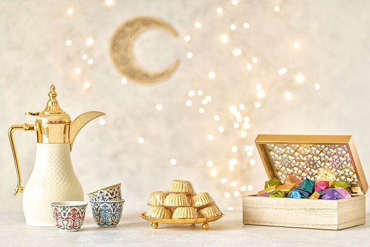 Ein Bild zum Ramadan für Muslime. Eine gold-weiße Kaffeekanne steht neben drei bunt gemusterten Tassen, die übereinandergestapelt sind. Daneben befindet sich ein Turm aus kleinen, goldfarbenen Gebäckstücken. Rechts steht eine geöffnete Holzschatulle, die mit bunten, glänzenden Pralinen gefüllt ist. Im Hintergrund sind eine goldene Mondsichel und zahlreiche kleine Lichtpunkte zu sehen.