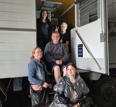 Fünf junge Menschen posieren vor einem weißen LKW mit offener Tür in einer Halle. Zwei Personen sitzen auf der Treppe zum Fahrzeug, eine Person sitzt vorne auf einem Hocker und trägt einen durchsichtigen Regencape. Zwei weitere Personen stehen seitlich und lächeln in die Kamera.