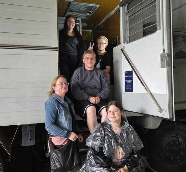 Fünf junge Menschen posieren vor einem weißen LKW mit offener Tür in einer Halle. Zwei Personen sitzen auf der Treppe zum Fahrzeug, eine Person sitzt vorne auf einem Hocker und trägt einen durchsichtigen Regencape. Zwei weitere Personen stehen seitlich und lächeln in die Kamera.