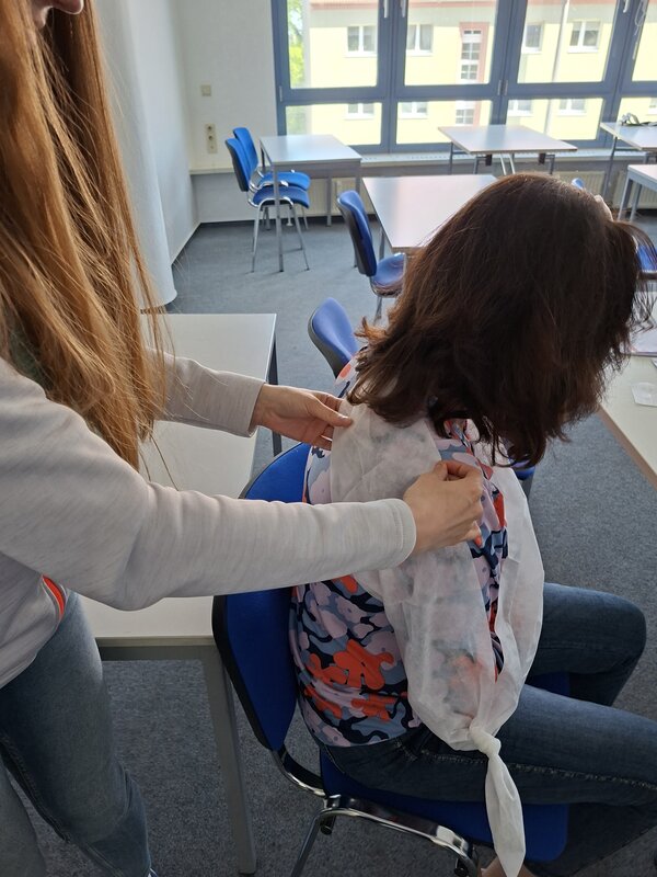 Eine Frau sitzt auf einem Stuhl und bekommt von einer anderen Frau einen Verband um die Schulter gelegt.