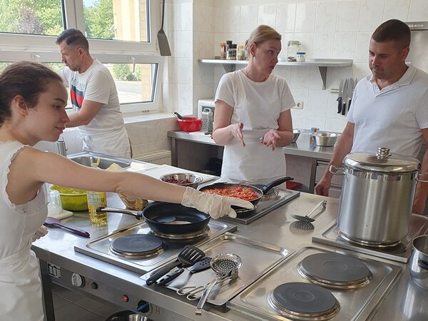 Eine Teilnehmerin erklärt einem Teilnehmer etwas am Herd. Zwei andere Teilnehmende bereiten weitere Speisen vor und zu.