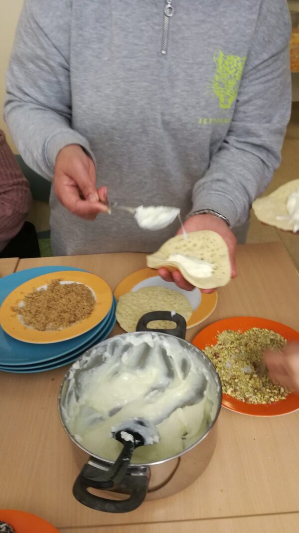 Eine Person in einem grauen Pullover hält einen Atayef in der Hand und füllt ihn mit einer weißen Creme. Auf dem Tisch liegen weitere Atayef, sowie Teller mit gehackten Nüssen und einem Topf mit Creme.