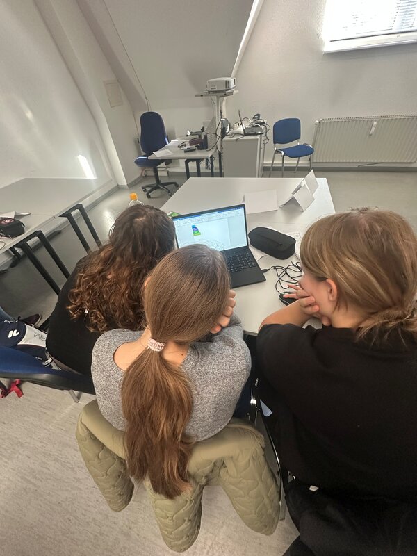 Drei Schülerinnen sitzen gemeinsam an einem Tisch und arbeiten an einem Laptop. Auf dem Bildschirm ist eine technische Zeichnung beziehungsweise eine Konstruktionssoftware geöffnet.