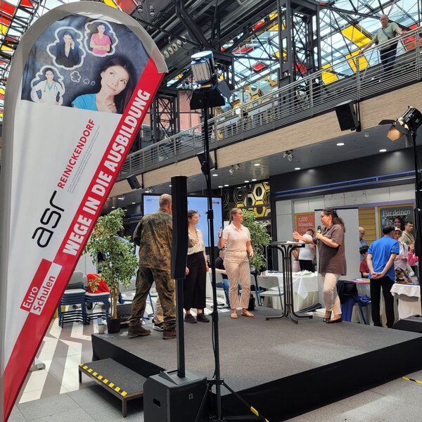 Die Vertreter*innen der beteiligten Ausbildungsunternehmen stellen auf der Bühne ihre Berufe vor. Links im Bild ist die Beach Flag der Euro-Schulen mit dem Schriftzug "ASR Reinickendorf - Wege in die Ausbildung" zu erkennen.