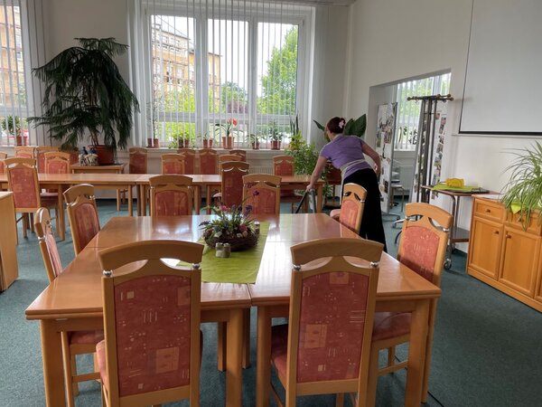 Ein großer Raum mit langen Holztischen und orange-rot bezogenen Stühlen. Im Vordergrund steht ein Tisch mit einer grünen Tischdecke und einem Blumengesteck. Eine Person in lila T-Shirt und schwarzer Hose saugt den Boden. Im Hintergrund sind große Fenster mit Pflanzen auf der Fensterbank sowie ein Whiteboard zu erkennen.