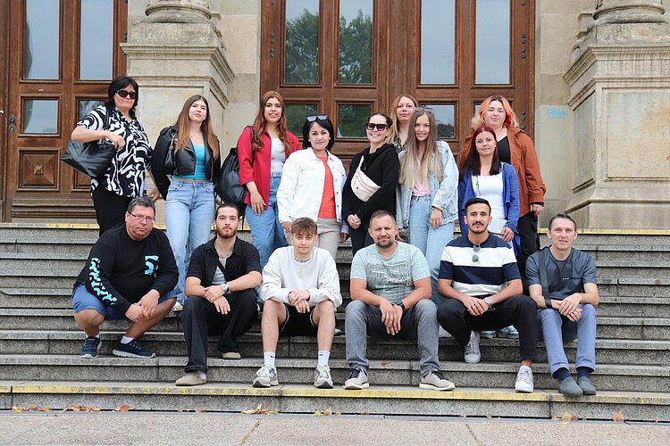 Unser Integrationskurs besucht Leipzig Gruppenfoto von 15 Personen auf einer Treppe vor einem historischen Gebäude mit Holztüren.
