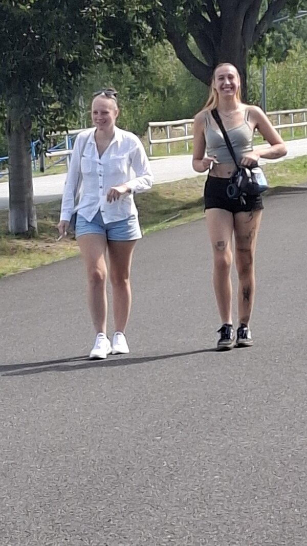 Zwei junge Frauen gehen bei sonnigem Wetter lächelnd auf einem von Bäumen gesäumten Weg entlang.