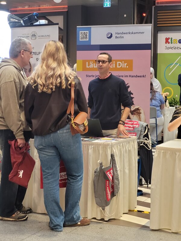 Ein junger Mann mit Brille steht hinter einem Messestand und spricht mit zwei Personen. Der Stand ist mit einem Roll-up der Handwerkskammer Berlin ausgestattet, auf dem ein QR-Code und der Slogan 'Läuft bei Dir. Starte jetzt deine Karriere im Handwerk!' zu sehen sind. Auf dem Tisch liegen Taschen und Infomaterialien. Im Hintergrund sind weitere Messestände und bunte Schilder sichtbar.