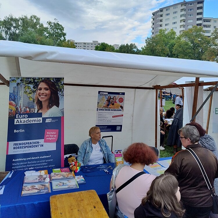 Mitarbeiterin der Euro-Schulen Berlin sitzt am überdeckten Stand und berät und unterhält sich mit Besucherinnen und Besuchern. Im Hintergrund steht ein großes Euro Akademie Roll-up, auf dem Tisch sind Informationsflyer und -broschüren ausgelegt.