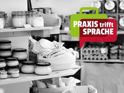 Ein Regal in einem Laden zeigt verschiedene Produkte wie Gläser mit Cremes, eingemachte Gläser und gefaltete Handtücher in Körben. Im Vordergrund ist eine grüne Sprechblase mit der Aufschrift 'PRAXIS trifft SPRACHE' in weißer und roter Schrift eingeblendet. Der Hintergrund des Bildes ist in Schwarz-Weiß gehalten, während die Sprechblase farbig hervorgehoben ist.