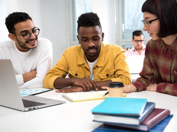 Zwei junge Männer und eine Frau sitzen an einem Tisch und schauen auf ein Notebook, davor liegen Bücher.