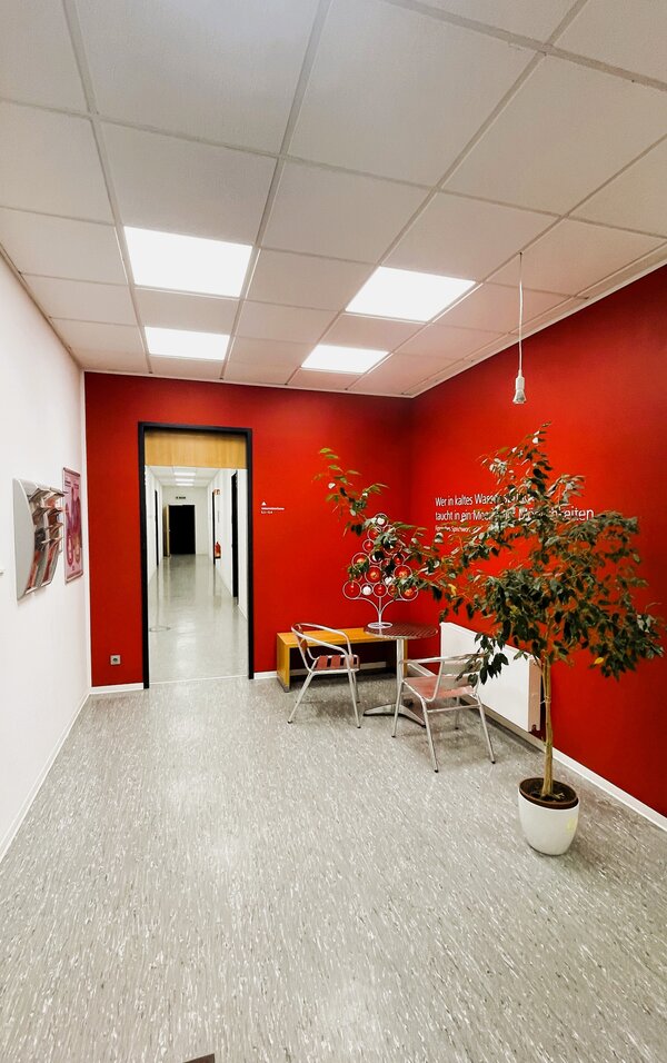 Sitznische mit roter Wand, kleinem Metalltisch und -stühlen, großem Zimmerbaum und weißem Kugel-Deko-Tannenbaum, im Hintergrund ein heller Flur mit Türen.