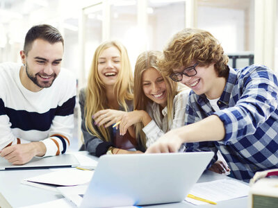 Vier junge Erwachsene sitzen an einem Tisch und arbeiten gemeinsam an einem Laptop. Die Person links, ein Mann mit Bart und einem gestreiften Pullover, lächelt. Die beiden Frauen in der Mitte, mit langen blonden Haaren, scheinen sich über etwas auf dem Laptop zu freuen. Der Mann rechts, mit Brille und einem karierten Hemd, zeigt auf den Laptop-Bildschirm. Auf dem Tisch liegen verschiedene Papiere und ein Stift.