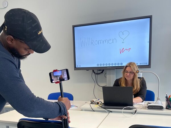 Mitarbeiterin sitzt vor dem Laptop am Schreibtisch und erklärt etwas, während der Kameramann sie aufnimmt. 