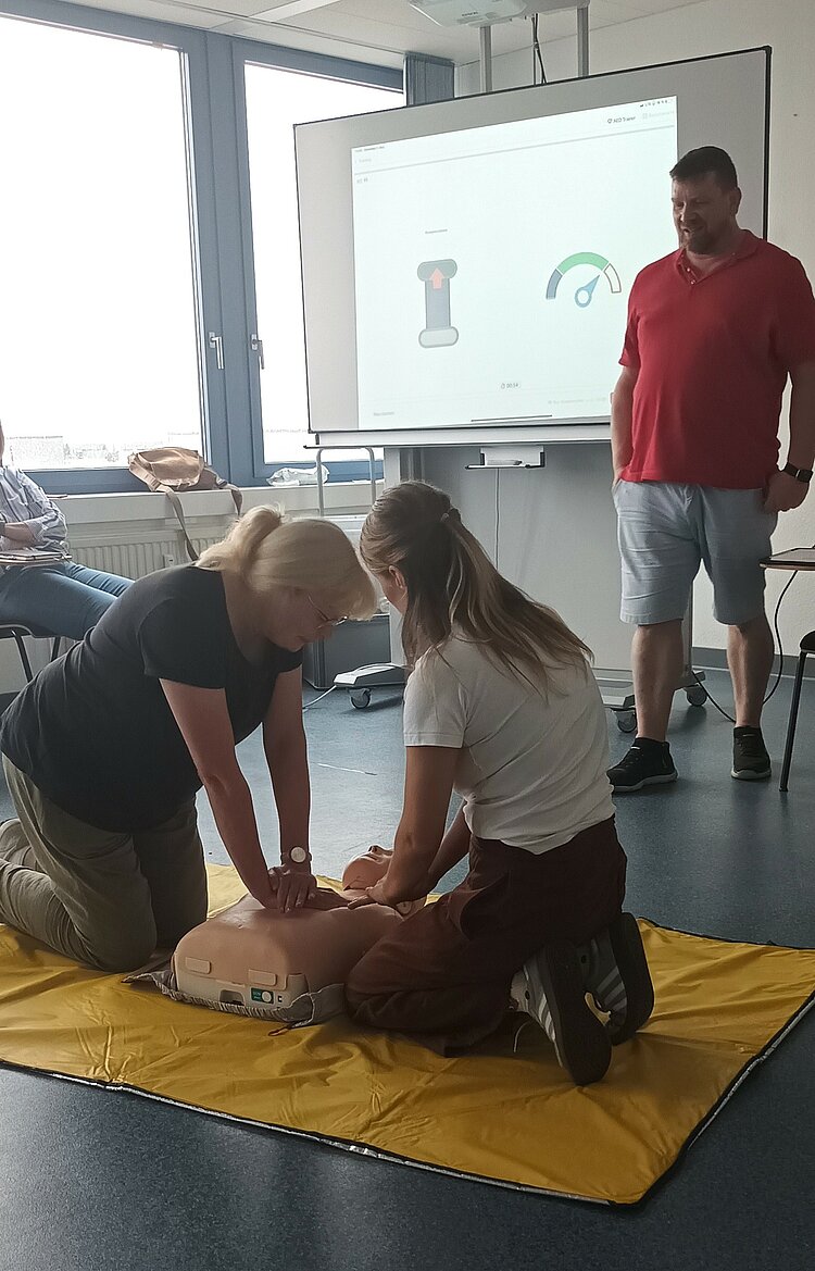 Zwei Personen üben Herz-Lungen-Wiederbelebung an einer Übungspuppe, während der Übungsleiter im Hintergrund steht und die Szene beobachtet. Die Übung findet in einem Raum mit einem großen Bildschirm statt, auf dem eine Präsentation zu sehen ist.