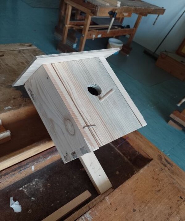 Ein handgefertigter Nistkasten aus hellem Holz steht auf einer Werkbank. Der Nistkasten hat ein geneigtes Dach, ein rundes Einflugloch und eine kleine Sitzstange darunter. Im Hintergrund sind Werkzeuge und Werkbänke in einer Werkstatt sichtbar.