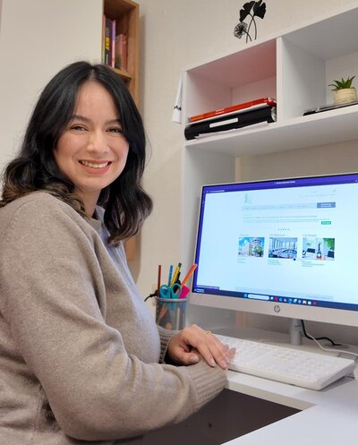 Eine weibliche Person mit schulterlangem, dunklem Haar sitzt an einem Schreibtisch vor einem weißen Computerbildschirm. Der Bildschirm zeigt eine Webseite mit mehreren Bildern und Textabschnitten. Auf dem Schreibtisch stehen ein weißes Tastatur, ein Becher mit bunten Stiften und ein Notizbuch. Im Hintergrund befinden sich weiße Regalfächer mit Büchern, einem geschlossenen Laptop und einer kleinen Pflanze. An der Wand ist eine schwarze Blumenillustration zu sehen.