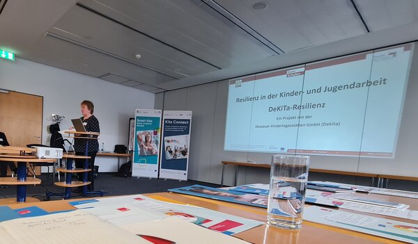 Foto eines Tagungsraums: Eine Referentin steht an einem Rednerpult vor einer Leinwand und hält einen Vortrag zum Thema „Resilienz in der Kinder- und Jugendarbeit – DeKiTa-Resilienz“. Auf der Folie ist der Projektpartner Dessauer Kindertagesstätten GmbH (Dekita) zu lesen, im Hintergrund stehen Roll-Ups von Euro-Schulen und Projektbannern, im Vordergrund liegen Tagungsunterlagen und ein Glas Wasser auf den Tischen der Teilnehmenden.
