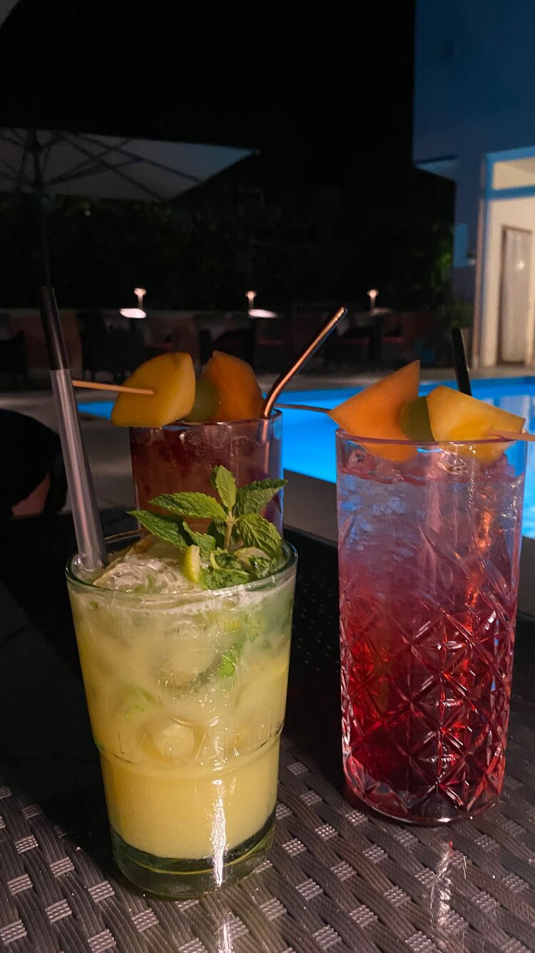 Auf einem Tisch neben einem beleuchteten Pool stehen drei unterschiedliche Cocktails, die mit Minze und Früchten dekoriert sind. Das Bild ist bei Nacht aufgenommen worden.