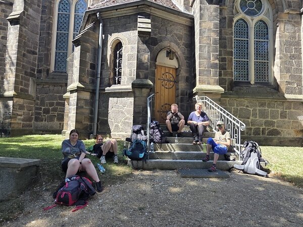 Eine Gruppe von Wanderern mit Rucksäcken sitzt entspannt auf den Stufen vor einer alten Steinkirche mit gotischen Fenstern und einer Holzür. Die Szene wirkt sonnig und gelöst.