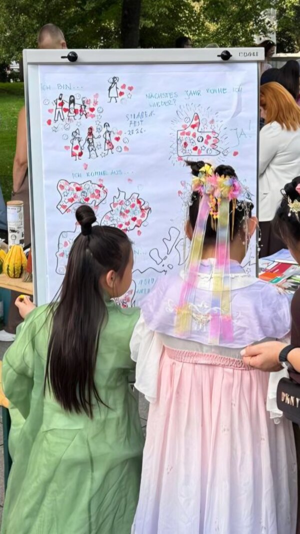 Vor einem großen Whiteboard mit handgezeichneten Illustrationen und handschriftlichen Notizen stehen zwei Mädchen in traditioneller Tracht; an einem Tisch dahinter liegen u. a. Kürbisse.