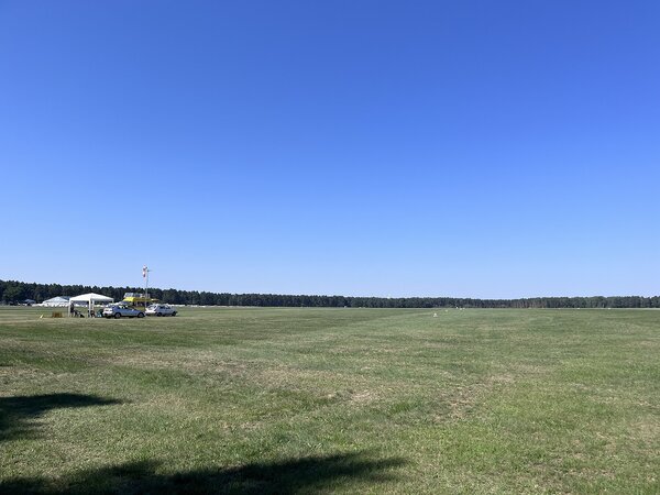 Weitläufiges Flugplatzgelände mit großer Rasenfläche unter blauem Himmel; am Rand stehen Fahrzeuge, Zelte und einige Personen.