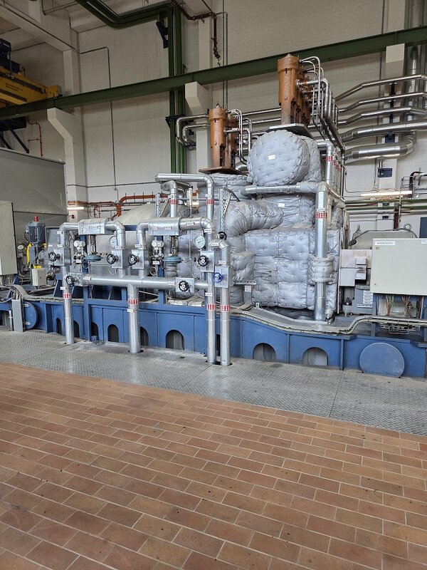 Technische Industrieanlage in einer Werkhalle mit zahlreichen Rohrleitungen, Armaturen und Messinstrumenten. Im Zentrum befindet sich eine große, mit grauem Isoliermaterial ummantelte Maschinenkomponente. Im Vordergrund verlaufen mehrere silberfarbene Rohrleitungen über einem blauen Maschinenfundament.
