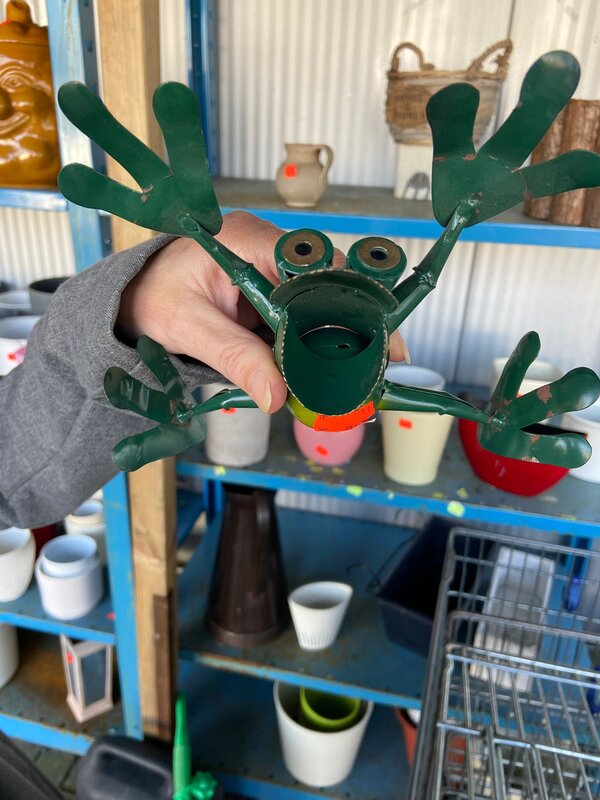 Eine Hand hält einen grünen Metallfrosch mit großen Augen und ausgestreckten Armen vor ein Regal mit Vasen, Töpfen und Dekoartikeln.