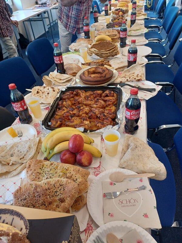 Lange gedeckte Tafel mit verschiedenen Brotsorten und Fladenbrot, einem großen Blech mit gebackenen Hähnchenteilen, Obst, Gebäck und Getränken in einem Seminarraum.