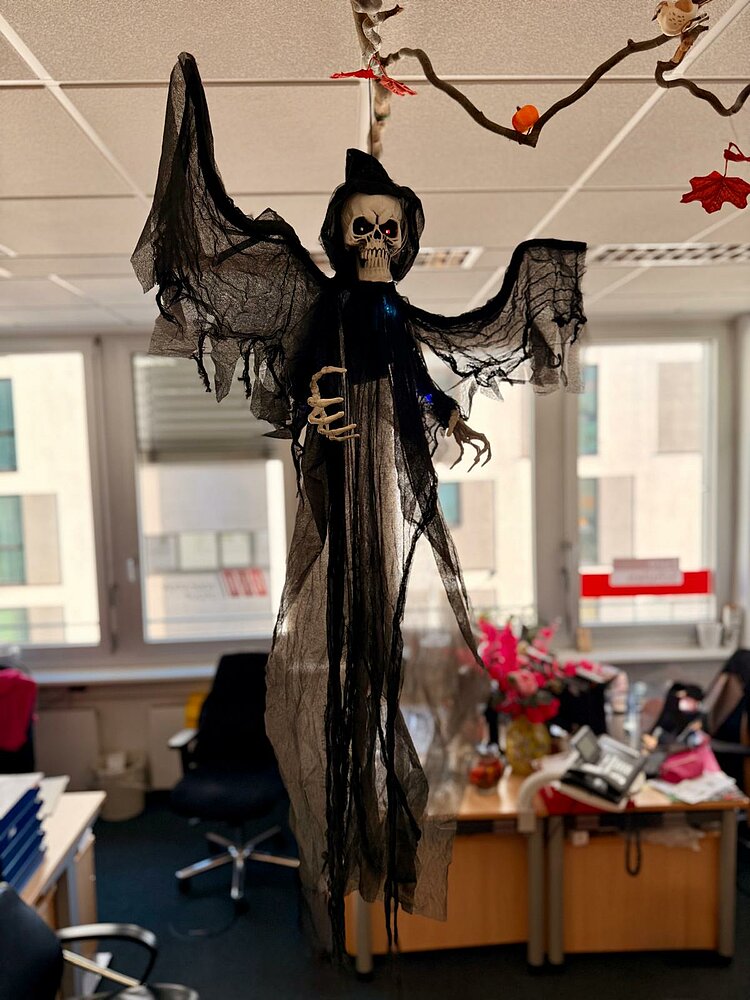 Eine gruselige Skelettfigur mit schwarzem, zerrissenem Umhang hängt von der Decke in einem Büro. Im Hintergrund sind Schreibtische, Stühle und Fenster zu sehen.