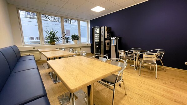 Aufenthaltsraum mit langer blauer Polsterbank, mehreren Holztischen, Metallstühlen, zwei Getränkeautomaten und großen Fenstern mit Blick nach draußen.