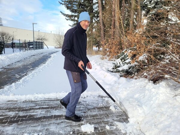 Hausmeister der Euro-Schulen Hohenstein-Ernstthal schippt der Schnee auf dem Gehweg mit einer Schneeschaufel weg. Er trägt eine Mütze und lächelt in die Kamera.