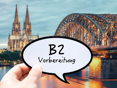 Eine Hand hält ein Sprechblasen-Schild mit der Aufschrift 'B2 Vorbereitung' vor einer Stadtlandschaft mit einem Fluss und historischen Gebäuden im Hintergrund. Die Szene scheint sich auf eine Vorbereitung oder einen Kurs zu beziehen, möglicherweise in einer deutschsprachigen Region. Im Hintergrund sind der Kölner Dom und die Hohenzollernbrücke zu sehen, was auf die Stadt Köln in Deutschland hinweist.
