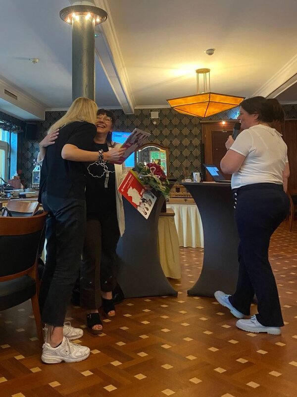 Drei Frauen stehen in einem Raum mit dekorativen Wandmustern. Eine Frau hält ein Mikrofon, eine andere hält ein Zertifikat und einen Blumenstrauß, während die dritte Frau sie umarmt.